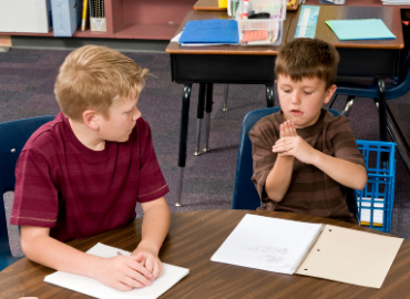 Two young boys at school, share a desk. The boy in the right hand is making a gesture, trying to explain something to his deskmate.