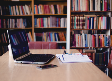 A laptop computer open, sitting on top a table, with bookshleves in the background